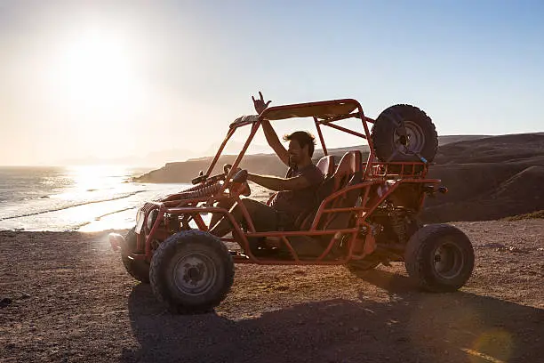 Active man driving quadbike on dirt road by the sea in sunset showing rocking sign to the camera.
