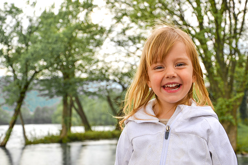 Portrait of a cute little girl with long blond hair, smiling with tounge sticking out of her mouth, wearing white jacket and having fun in the nature by the river; Photo focused on girl's cute smile with green trees and river in the background.