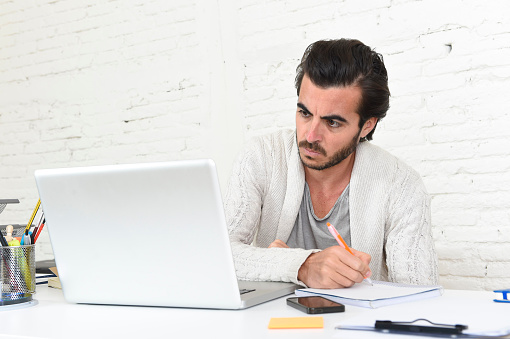 young attractive student preparing university project writing on pad with pen or hipster style freelancer businessman working with laptop computer analyzing project at home office studio