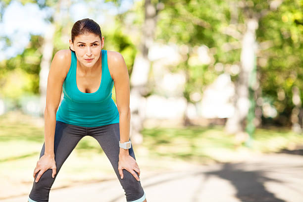 Exhausted Female Jogger At Park Tired young female jogger standing with hands on knees after run in park. Horizontal shot. hand on knee stock pictures, royalty-free photos & images