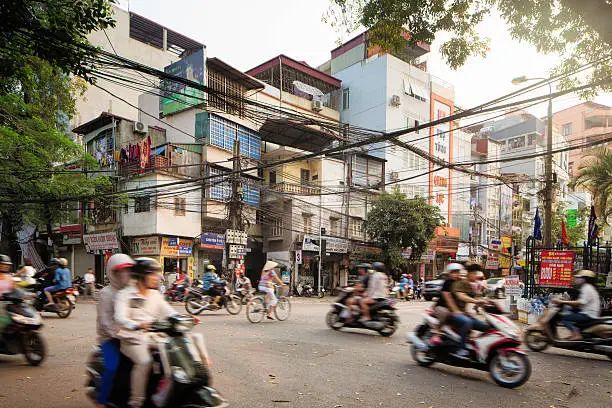 Morning Traffic on Lo Duc Street in Hanoi, Vietnam on a warm Spring day. People are commuting on bikes and motor bikes, or walking and shopping. Apartment buildins are rising above the street behind chaotic electric cables.