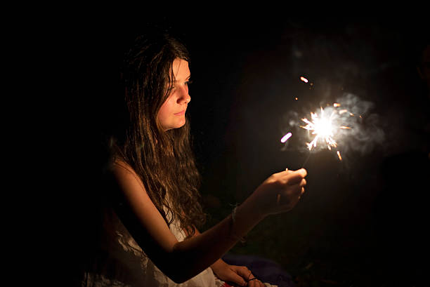 A girl holds a sparkler up, she is lit by the sparkler.  Behind her is all black, but we can see her face adn hair and arm holding the sparkle