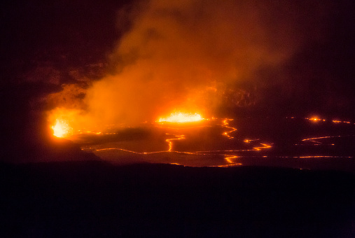Orange molten magma erupts at night inside Hawaii Island's Kilauea volcano. The Lava lake is glowing orange inside a volcanic caldera. A cloud of volcanic gasses is illuminated by glowing molten lava splattering into the air. The night image is horizontal and mostly dark except for the glowing areas.