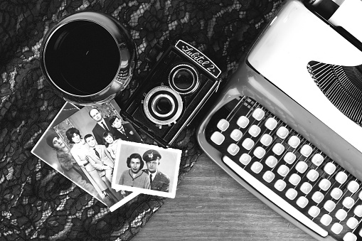 Konak, Izmir, Turkey- March 12, 2016: A wineglass ,typewriter, old Lubitel 2 analog twin -lens reflex camera and old black and white photos are on the wooden table with lace textile. Small photo is about a wife and a husband who wears soldier uniform. Second photo which is staying at the middle is about a family. And family includes a stylish wife and husband and three children. A small boy at the right side of the photo wears a school uniform. Third photo at the under of the all photos is about a stylish woman who wears a hat and a fur coat.