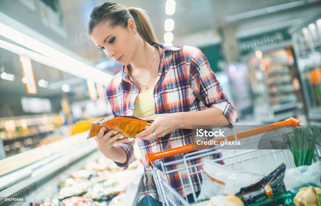 Woman buying food in supermarket. Closeup of mid 20's blond woman leaning over large freezer in a supermarket and reaching for some frozen food. She's carrying red shopping basket which is almost full of unrecognizable items. Tilt shot. Supermarket Stock Photo
