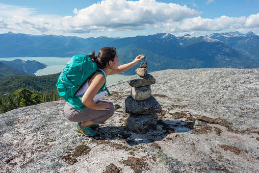 A young, female Asian backpacker adds a stone to a cairn on a mountaintop in Squamish, British Columbia, Canada.  View of the valley and surrounding mountains in the background.