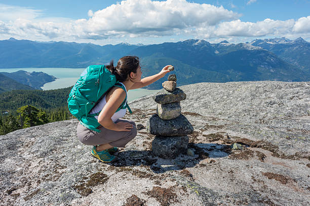 Young Woman Hiker Placing Rock on Cairn on Mountain Summit A young, female Asian backpacker adds a stone to a cairn on a mountaintop in Squamish, British Columbia, Canada.  View of the valley and surrounding mountains in the background. cairn stock pictures, royalty-free photos & images
