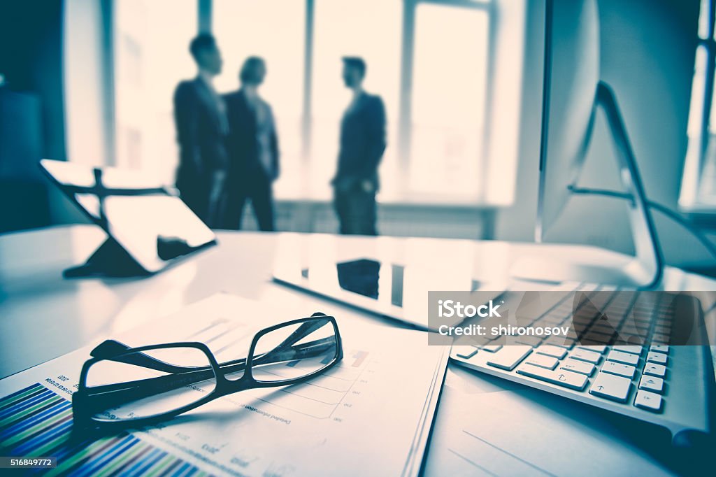 Business workplace Modern devices, documents, eyeglasses on the foreground, unrecognized businessmen on the background  Adult Stock Photo