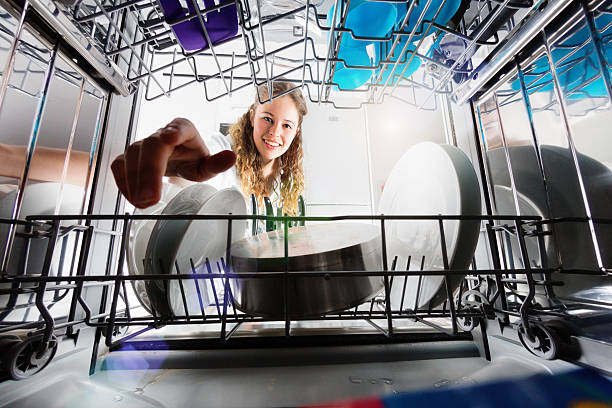 Seen from inside dishwasher, cute smiling girl loading or unloading Seen, unusually, from inside the dishwasher drum, a pretty blonde teenager smiles as she loads ir unloads dishes. plate rack stock pictures, royalty-free photos & images