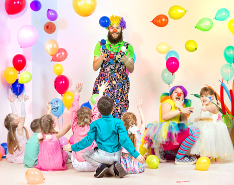 The group of well-dressed children of preschool and elementary age are having fun at holiday. Two cheerful clowns are entertaining the kids at a children's party. The bearded clown juggling colorful balls. Children are watching a clown performance. Cute female clown is painting on girl's face. Shooting at studio among colorful balloons
