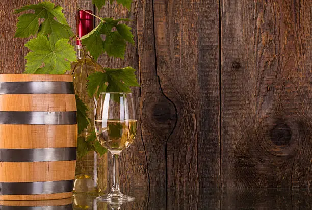Glass of wine with barrel white bottle behind grapeleaves and dark wooden background