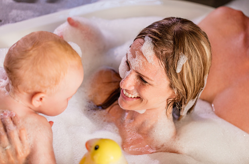 Close up of mom and daughter playing in bubble bath with soap suds and father sitting in bath behind mother