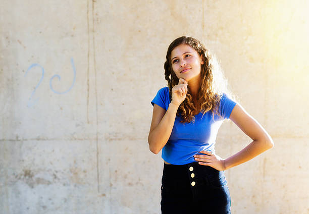 This beautiful young honey-blonde woman with one hand on her hip, the other on her chin as she thinks, smiles gently as she stands next to a blank space - ready for your message - on a grungy grey wall, lit by sunshine.