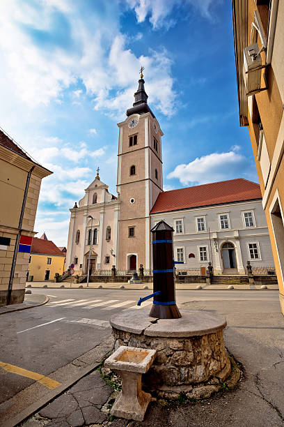 A Baroque-style church with an ornate facade and clock tower stands in a town square. The church is surrounded by other historic buildings and parked cars, under a cloudy sky, highlighting the town's rich architectural heritage.