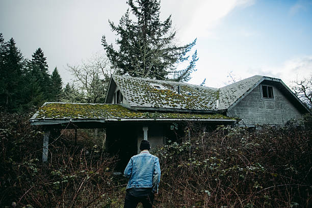 Real lifestyle photo of a young male explorer walking towards an abandoned home in the middle of a forest. This home was clearly abandoned for many years and has plants growing all over the inside and out of it. This image shows the young man walking towards the house interested in seeing what might be inside.