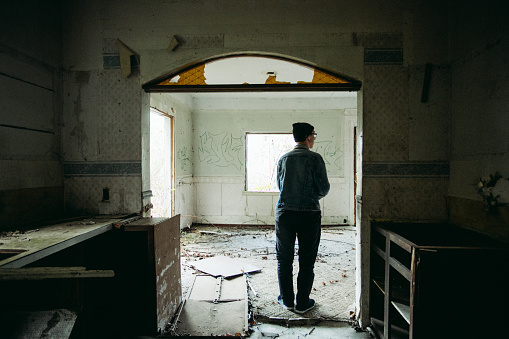 Real lifestyle photo of a young male explorer walking around inside an abandoned home in the middle of a forest. This home was clearly abandoned for many years and has been trashed by squatters over the years. This image shows the young man walking inside the house interested in seeing what might be around the corner.