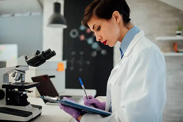 Female researcher taking notices at her workplace. She is standing in her laboratory, using microscope and laptop for her research.