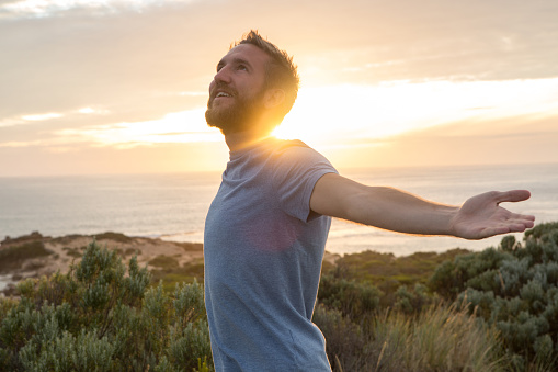 Cheerful young man arms outstretched at sunset, coastline on the background.