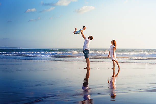 Happy family walking with fun on sunset sea beach Happy family - father, mother, baby son walk with fun along edge of sunset sea surf on black sand beach. Active parents and people outdoor activity on summer vacations with children on Bali island raro stock pictures, royalty-free photos & images