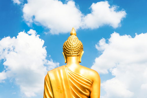 Golden Buddha statue from back focused on head  on white cloudy blue sky in sunny day