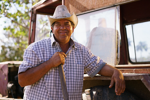 Farming and cultivations in Latin America. Portrait of middle aged hispanic farmer sitting proud in his tractor at sunset, holding the volante. He looks at the camera and smiles happy.