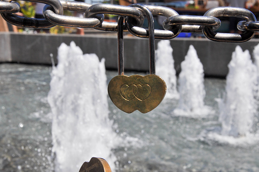 Closeup detail of gold etched love locks in the shape of a double love heart hanging from chain link with a blurred water fountain background outside the Bell Tower in Perth, Western Australia.