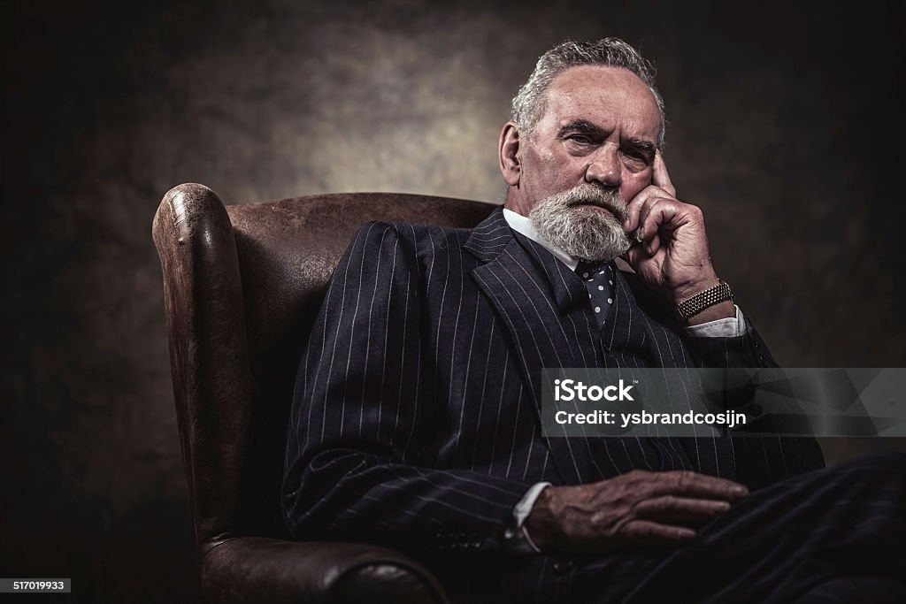 In chair sitting characteristic senior business man. Gray hair. In chair sitting characteristic senior business man. Gray hair and beard wearing blue striped suit and tie. Against brown wall. Contemplation Stock Photo