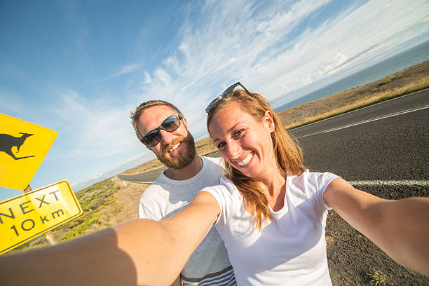 Young couple take selfie portrait near kangaroo warning sign-Australia Cheering young couple take a selfie portrait on the road standing next to a kangaroo warning sign, Australia. kangaroo crossing sign stock pictures, royalty-free photos & images
