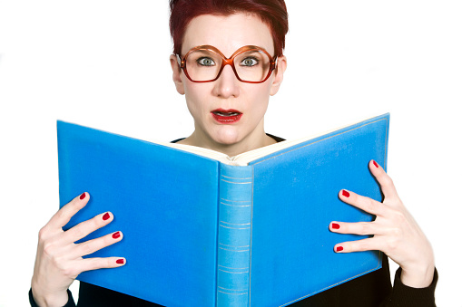 red-haired woman with glasses reading in a blue book and looking surprised