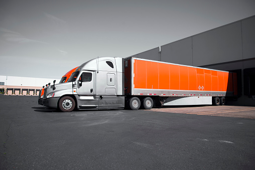 Two modern professional commercial semi trucks in gray and orange with dry van trailers are standing in the dock on territory of the transport warehouses for loading or unloading of commercial cargo for transportation over long distances.