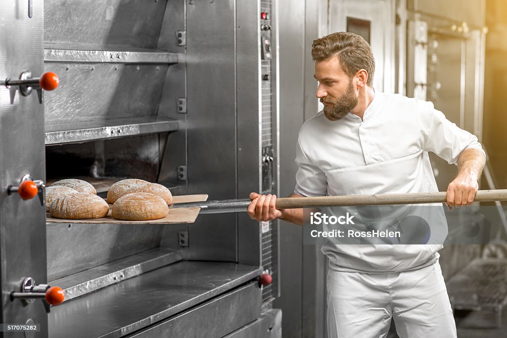 Baker taking out from the oven baked buckweat bread Handsome baker in uniform taking out with shovel freshly baked buckweat bread from the oven at the manufacturing Bread Stock Photo