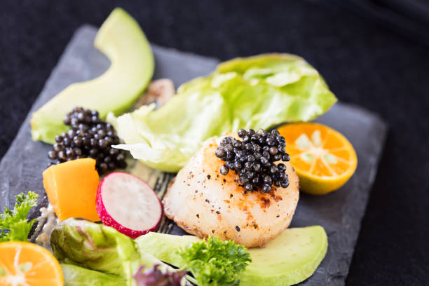 Pan seared scallops with salad, avocado, radish, mango and black caviar on a stone plate.