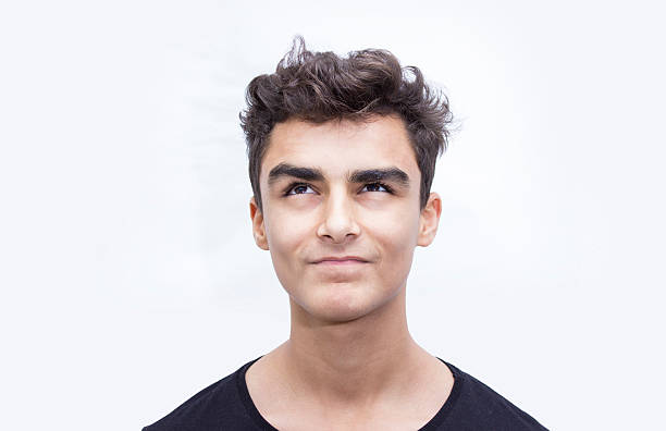Front view portrait of smiling child thinking over white background. Young man wearing casual clothes. Thinking boy standing in front of isolated on white and looking up with smiling facial expression. Horizontal compositon. Teenage boy's ethnicity belongs to Turkish, middle eastern ethnicity. Close-up shot. Young boy has got short, brown hair.