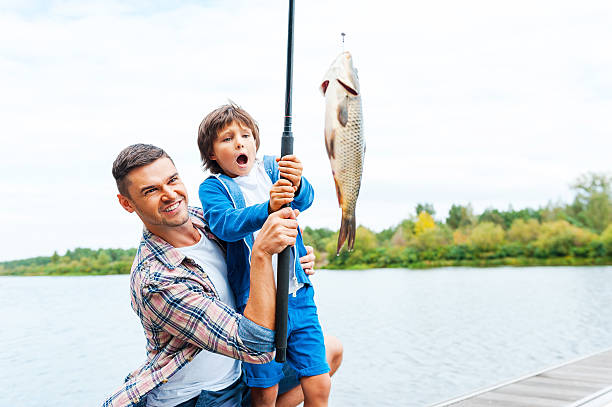 It is so big! Father and son stretching a fishing rod with fish on the hook while little boy looking excited and keeping mouth open fishing rod photos stock pictures, royalty-free photos & images