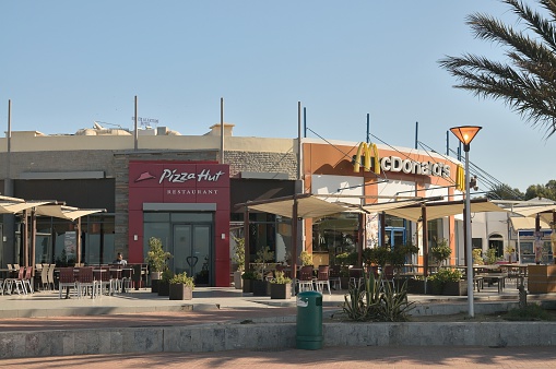 Agadir, Morocco - February 17, 2016: McDonalds and Pizza Hut fast food restaurants in Agadir, Morocco along the Plage D'Agadir with a lone Berber Moroccan man sitting at an outdoor table.