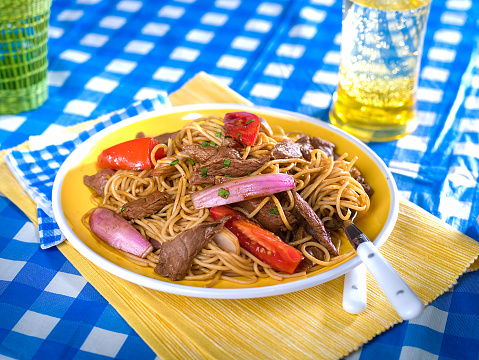 Beef and noodle stir fry, tallarin saltado, a typical Peruvian dish served on yellow plate in picnic setting with blue and white checked table cloth