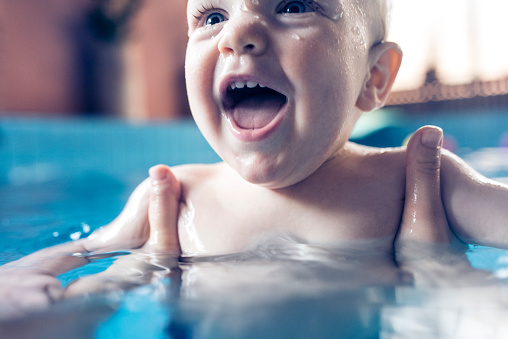 Nine month old baby boy at his first swimming lesson. Smiling and enjoying water. Mother is holding him and helping him to swim.