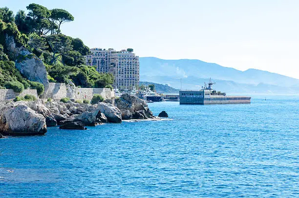 The rugged coastline of the Cote d'Azur on approach to the port of Cap d'Ail which neighbours Monaco. Trees cling to a hillside overlooking the rocky coastline.