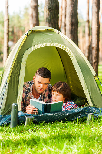 Father and son reading book while lying in tent together