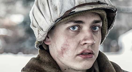 Close-up portrait of a serious young WWII US Army combat infantry soldier looking slightly sideways at the camera. His face is very cold with ruddy red cheeks, a bit of frostbite on his upper lip, and deep blue eyes. He's wearing a white cloth covering as camouflage on his standard issue metal helmet on top of an authentic US WWII military uniform knitted skull cap. The soldiers in this photo series are dressed in complete, completely period-correct uniforms with historically accurate weapons, helmets, supplies, equipment, tools, etc. Professional reenactment actors/models at the January-February 2016 Utah 