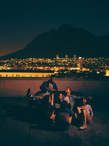 Group of multi-ethnical young hipster friends partying around a couch on a rooftop party. Citylights and night skyline in the background.