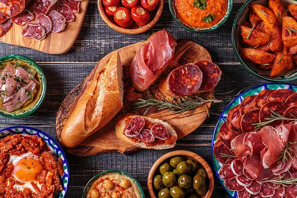 Typical spanish tapas concept. Concept include variety slices jamon,  chorizo, salami, bowls with olives,  peppers, anchovies, spicy potatoes, mashed chickpeas on a wooden table.