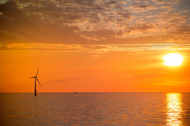 Wind turbine and lone boat in a calm ocean as the sun sets in the distance under a stunningly beautiful cloudscape.