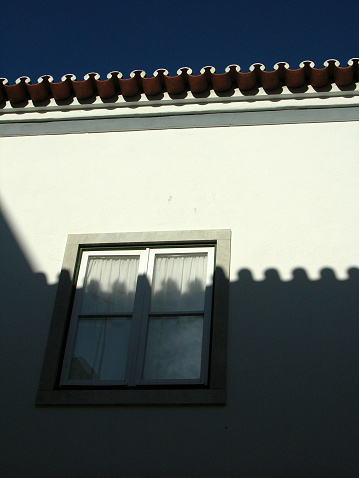View of closed window on façade of building with roof in tile of half cane typically Portuguese, with shadow of the adjacent roof.