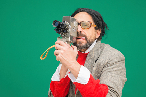 An untidy bizarre man, wearing big glasses, filming a home movie with an antique wind-up super 8 camera, over green background