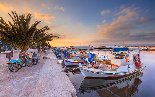 Greek fishing harbor scene with boats, palm trees and scooters at sunrise on a beautiful tranquil summer day in july