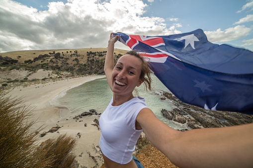 Cheerful young woman stands on a cliff above a beach in Kangaroo Island holding an Australian's flag.