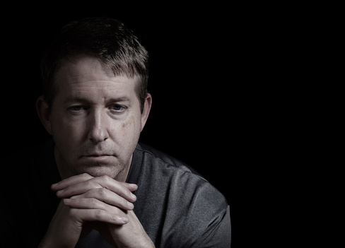 Closeup front view of mature man, looking forward, with chin resting in folded hands on dark background