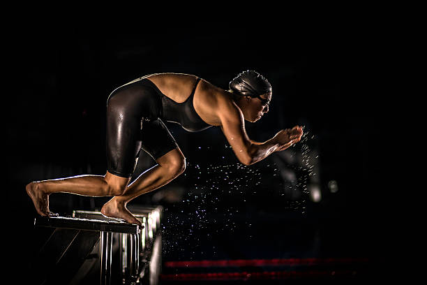 Female Swimmer Jumping Off The Starting Block Side view of young female at starting breaststroke race. black women in bathing suits stock pictures, royalty-free photos & images