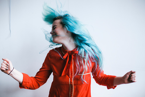 Portrait of a young woman with vibrant blue hair spinning around and head banging. Shot in a studio on a white background with 50mm lens, using all natural light.